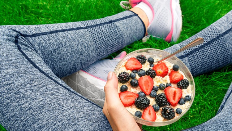 Person in grauer Sportkleidung sitzt auf einer Wiese und hält eine Schale mit Haferflocken, Erdbeeren, Blaubeeren und Brombeeren.
