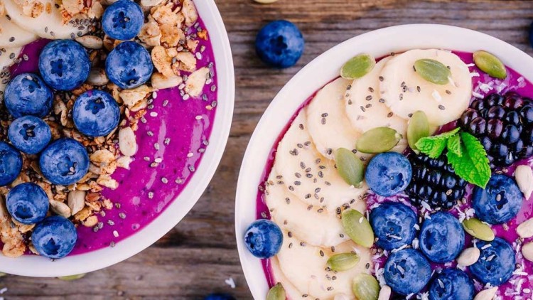 Zwei Schalen mit lila Smoothie, Banane, Heidelbeeren, Brombeeren, Körnern, Minzblättern und Granola auf Holztisch.