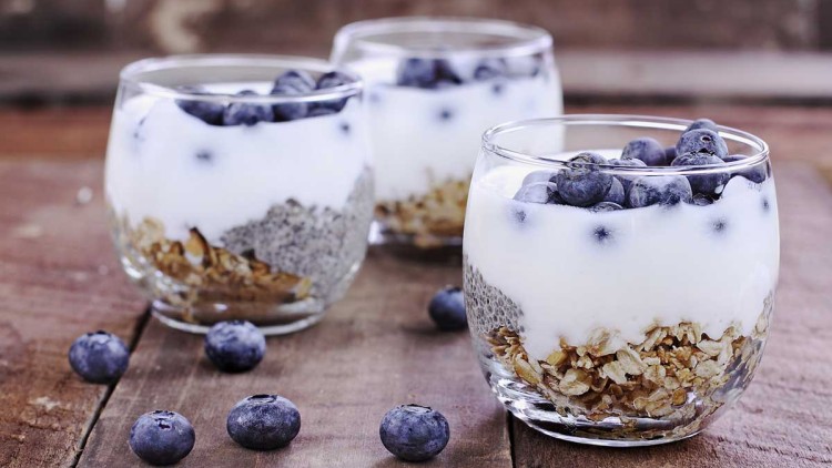 Drei Gläser mit Joghurt, Haferflocken, Chiasamen und frischen Heidelbeeren, auf Holztisch, mit verstreuten Heidelbeeren.