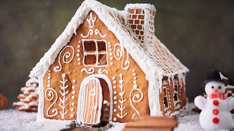 Ein Lebkuchenhaus mit weißem Zuckerguss und filigranen Verzierungen steht auf künstlichem Schnee, daneben ein Schneemann.