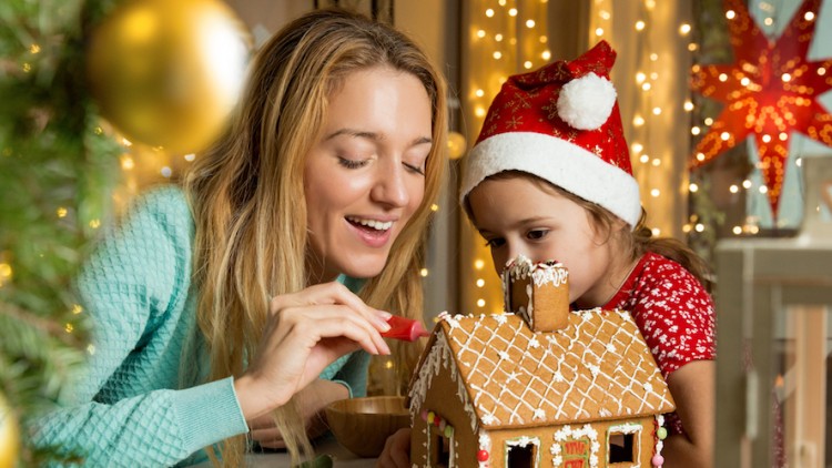 Person dekoriert ein Lebkuchenhaus mit Zuckerguss, im Hintergrund Lichterketten, Weihnachtsbaum und ein roter Stern.