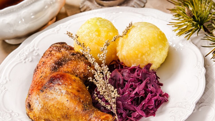 Gebratene Entenkeule mit zwei Kartoffelklößen, Rotkohl und Kräutergarnitur auf weißem Porzellanteller, im Hintergrund Soßenschale.