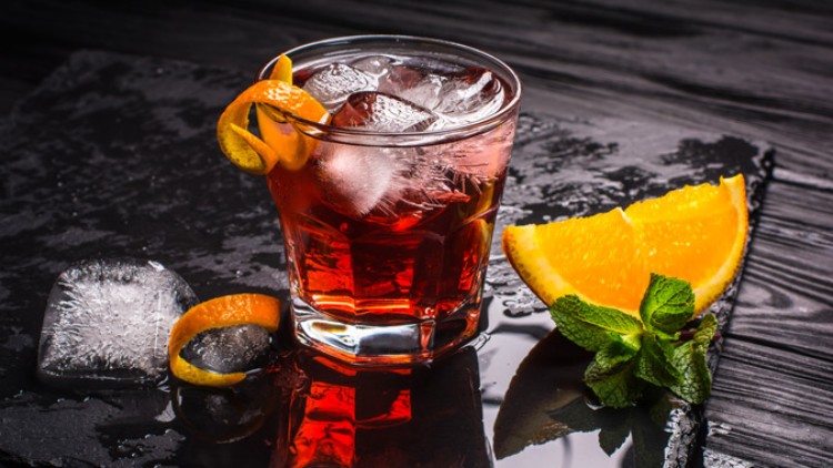Ein mit Eiswürfeln gefülltes Glas mit rotem Getränk, Orangenscheibe, Orangenschale und Minzzweig auf dunklem Untergrund.