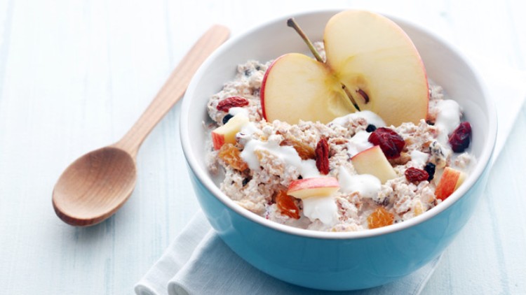 Eine blaue Schale mit Müsli, Joghurt, Apfelstücken, getrockneten Beeren und einer Apfelhälfte, daneben ein Holzlöffel.