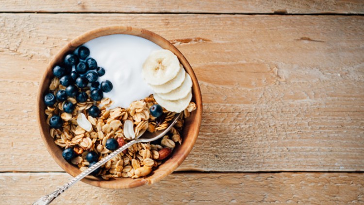 Eine Holzschale mit Joghurt, Müsli, Heidelbeeren, Bananenscheiben und einem silbernen Löffel steht auf einer hellen Holzfläche.