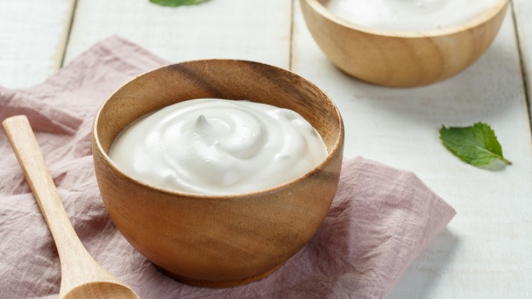 Holzschale mit cremigem weißen Joghurt, daneben ein Holzlöffel auf einem rosa Stofftuch, im Hintergrund grüne Blätter.