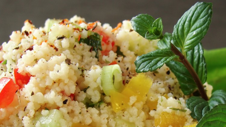 Nahaufnahme von Couscous-Salat mit roten, gelben und grünen Gemüsestücken, dekoriert mit frischen Minzblättern auf Salat.