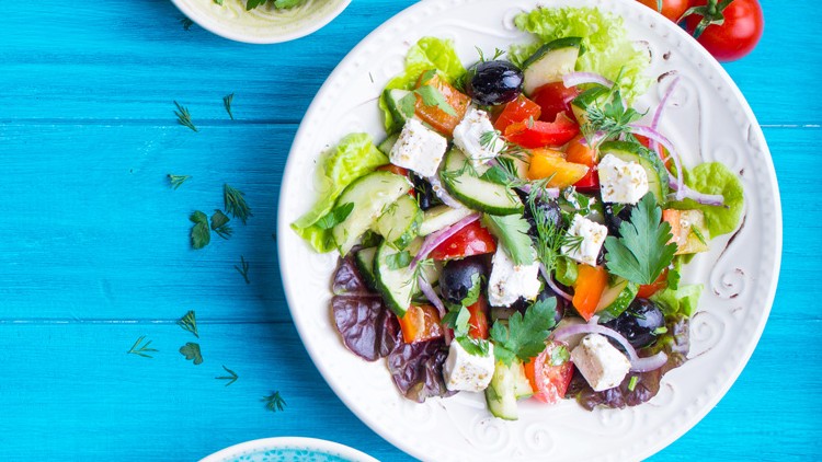 Weißer Teller mit gemischtem Salat aus Gemüse, Feta und Oliven, daneben Kräuter, Tomaten und eine kleine Schale auf blauem Untergrund.