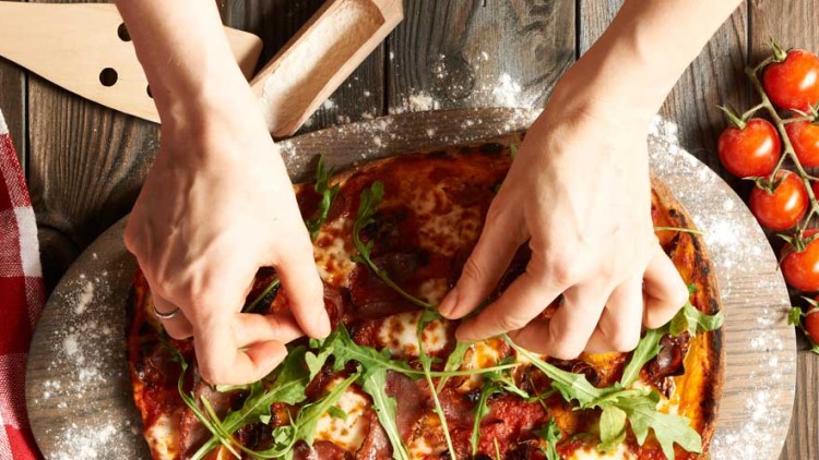 Hände legen frische Rucolablätter auf eine fertig gebackene Pizza auf einem bemehlten Holzbrett, daneben Kirschtomaten und ein Holzheber.