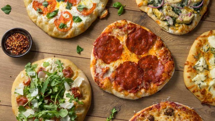 Sechs verschiedene Pizzen mit Belägen wie Salami, Tomaten, Rucola, Käse und Gemüse, auf einem Holztisch angerichtet.
