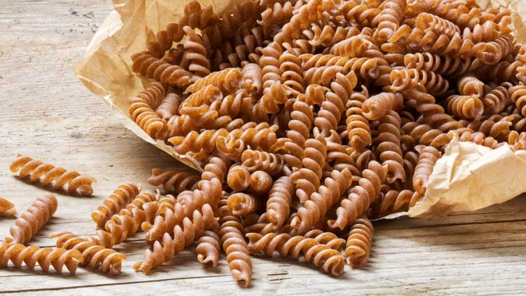 Braune Spiralnudeln aus Vollkorn liegen teils lose, teils in einem aufgerissenen Papierbeutel auf einer hellen Holzoberfläche.