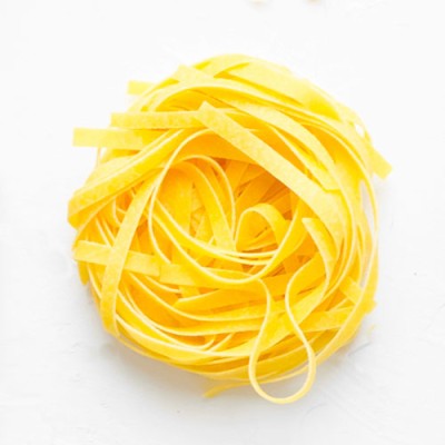 Ein Nest aus gelben, flachen Bandnudeln liegt auf einer weißen, strukturierten Oberfläche.