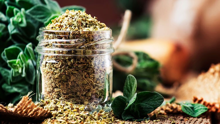 Ein Glas mit getrocknetem Oregano steht auf einem Tisch, umgeben von frischen Oreganoblättern und natürlichen Dekorationen.