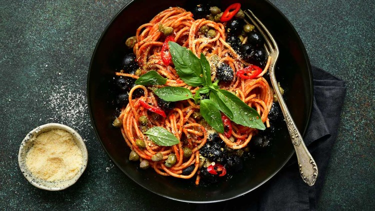 Ein Teller mit Spaghetti, Tomatensauce, schwarzen Oliven, Kapern, roten Chilistreifen und Basilikumblättern, daneben Parmesan.