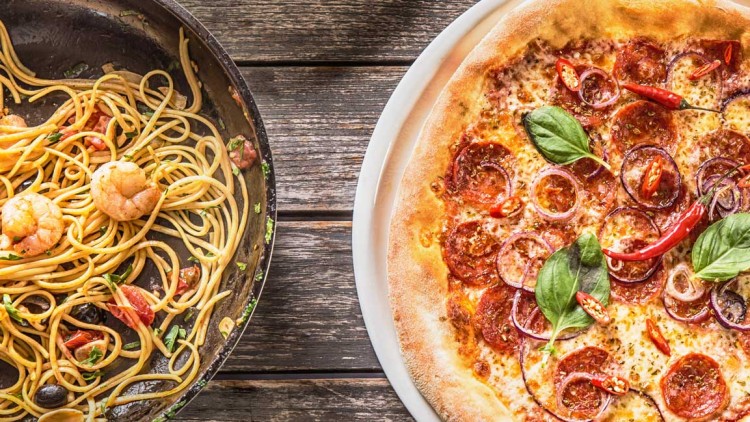 Spaghetti mit Garnelen in einer schwarzen Pfanne, daneben Pizza mit Salami und Chili, Süßkartoffelchips und roter Chili auf Holz.