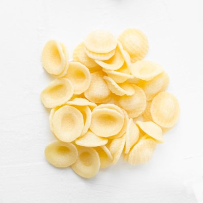 Gelbliche, ungekochte Orecchiette-Nudeln liegen locker auf einer weißen Oberfläche.