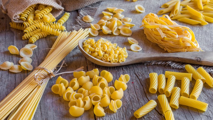 Verschiedene Sorten ungekochter Nudeln wie Penne, Spaghetti und Fusilli liegen auf einem Holztisch und Jutestoff.
