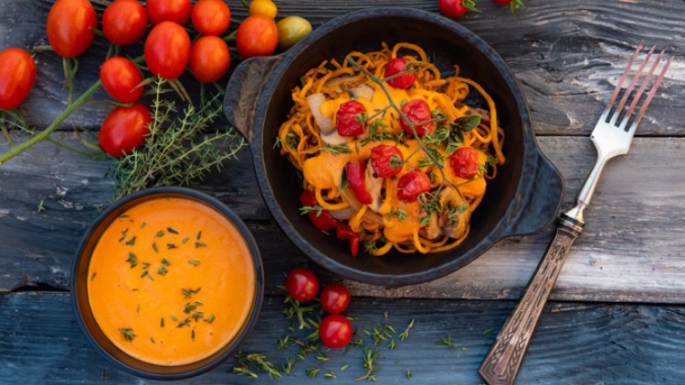 Ein schwarzer Teller mit Gemüsenudeln, überbacken mit Käse und Tomaten, daneben ein Schälchen Tomatensuppe und frische Kräuter.