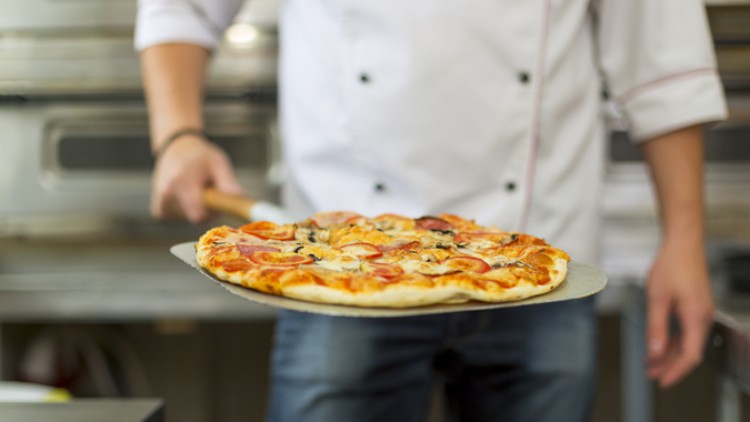 Eine Person in Kochkleidung hält eine Pizza mit Belag wie Tomaten, Käse und Oliven auf einem Backblech in einer Küche.