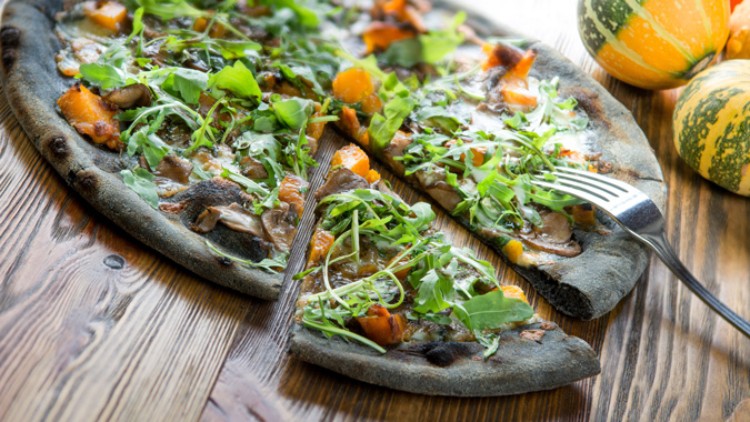 Eine dunkle Pizza mit Gemüse, Rucola und Pilzen liegt auf einem Holztisch, ein Stück ist herausgeschnitten, daneben Kürbisse.