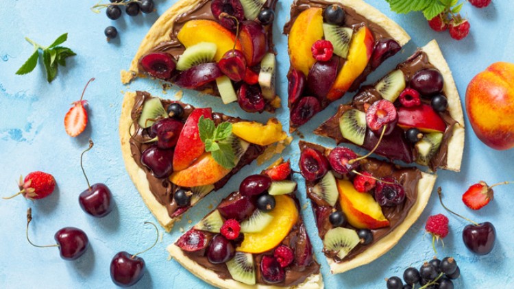 Runder Obstkuchen mit Schokocreme, belegt mit Kirschen, Kiwi, Pfirsich, Beeren und Minze, auf blauem Untergrund.