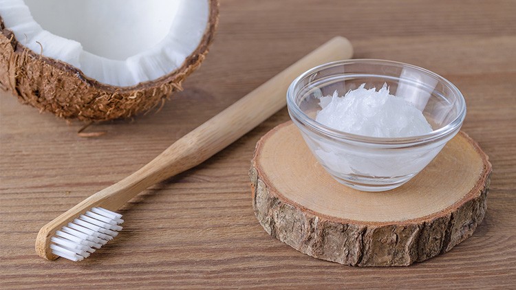 Eine geöffnete Kokosnusshälfte, ein Bambuszahnbürstengriff und ein Glasschälchen mit Kokosöl auf einer Holzscheibe.