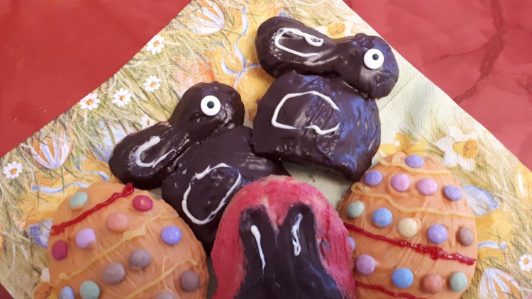 Schokoladenhasen und bunte Kekse in Eiform mit Glasur und Zuckerstreuseln auf einer Serviette mit Frühlingsmotiven.