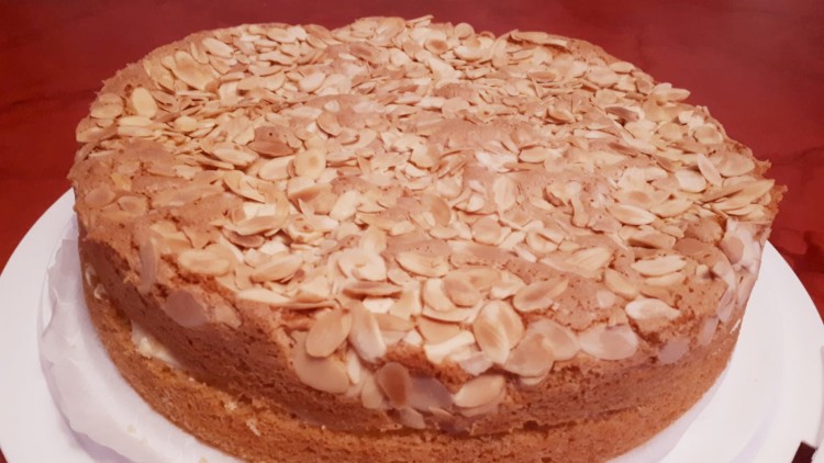 Runder Bienenstich-Kuchen mit einer goldbraunen Kruste aus Mandelblättern auf weißer Servierplatte.