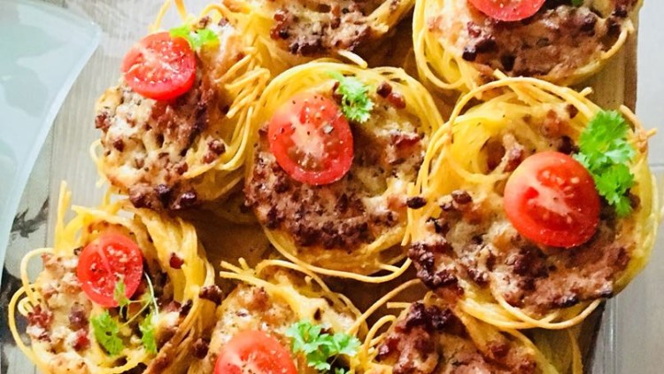 Mehrere gebackene Nester aus Spaghetti auf Holzplatte mit Hackfleisch, halben Tomaten und grüner Petersilie garniert.