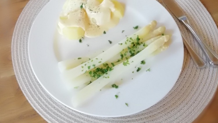 Tisch mit weißem Teller, gekochten Kartoffeln, weißem Spargel und Kräutern, daneben ein Weinglas, Besteck und eine Weinflasche.
