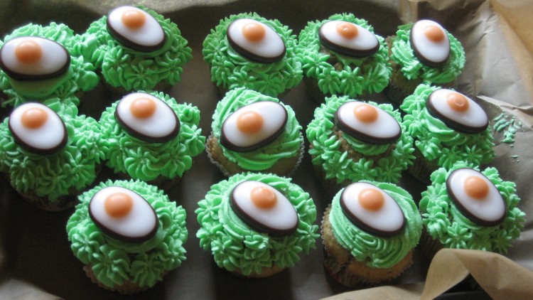 Zwölf Muffins mit grüner Creme und Dekoration aus Schokolade, weißem Guss und einer orangefarbenen Kugel auf Backpapier.