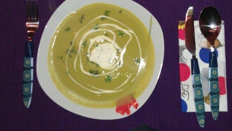 Ein weißer Teller mit gelber Suppe, Sahne und Kräutern steht auf lila Platzset, daneben Besteck, Glas und Serviette.