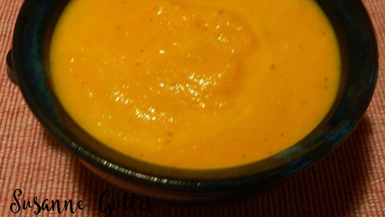 Eine dunkelblaue Schale mit orangefarbener Suppe steht auf einer roten Stoffunterlage, Name „Susanne Güttler“ ist unten links.