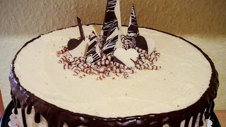 Runde Sahnetorte mit Schokoladenglasur am Rand und Schokoladendekoration in der Mitte auf schwarzer Tortenplatte.