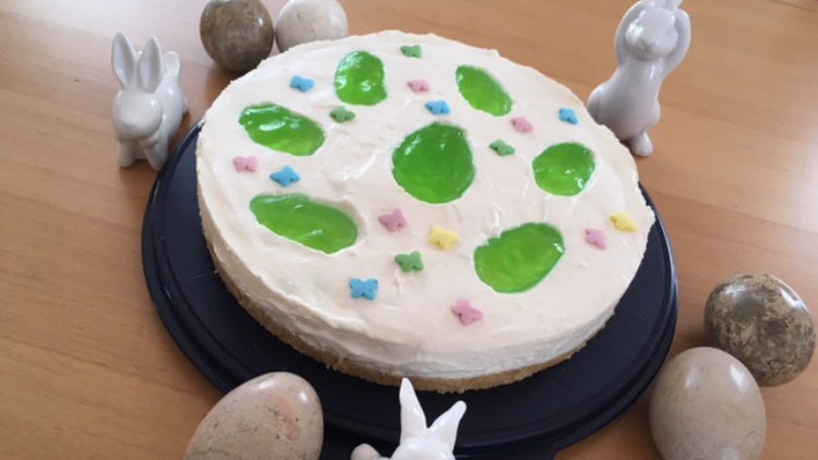 Weiße Torte mit grüner Gelatine und bunten Zuckerdekoren, umgeben von Keramik-Osterhasen und Steineiern auf Holztisch.