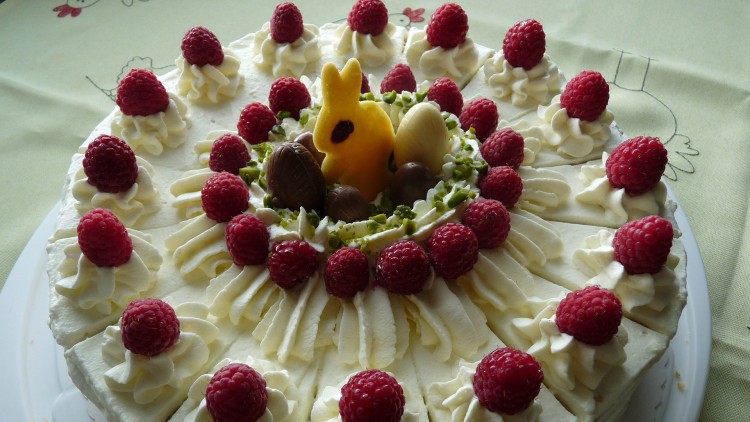Runde Sahnetorte mit Himbeeren, Sahnetupfen und Schokoladendekoration auf einem weißen Teller, in der Mitte ein gelbes Häschen.