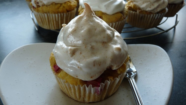 Ein Cupcake mit hellbraunem Baiser-Topping auf einem Teller mit Löffel, weitere Cupcakes im Hintergrund auf einem Gitter.