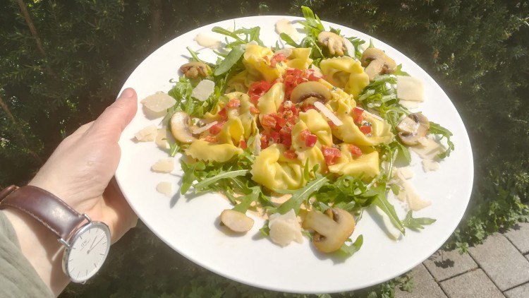 Weißer Teller mit gefüllten Tortellini, Rucola, geschnittenen Champignons, roten Paprikawürfeln und Parmesanspänen, im Freien gehalten.