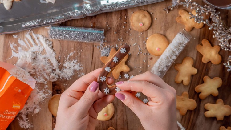 Hände dekorieren einen Schokokeks mit weißen Schneeflocken, umgeben von Backzutaten, Keksen und Deko auf Holztisch.