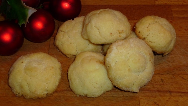 Mehrere helle Kekse auf einem Holztisch, dekoriert mit roten Weihnachtskugeln und einem Zweig mit grünen Blättern.
