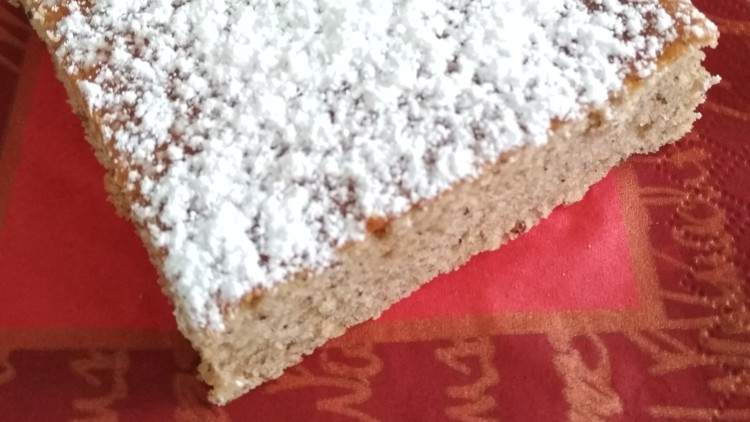 Ein rechteckiges Stück Kuchen mit Puderzucker liegt auf einer roten Serviette auf einem weißen Teller.
