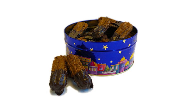 Blaue Keksdose mit Sternenmuster und bunten Häusern gefüllt mit braunen, schokolierten Spritzgebäck-Stücken, einige davor.