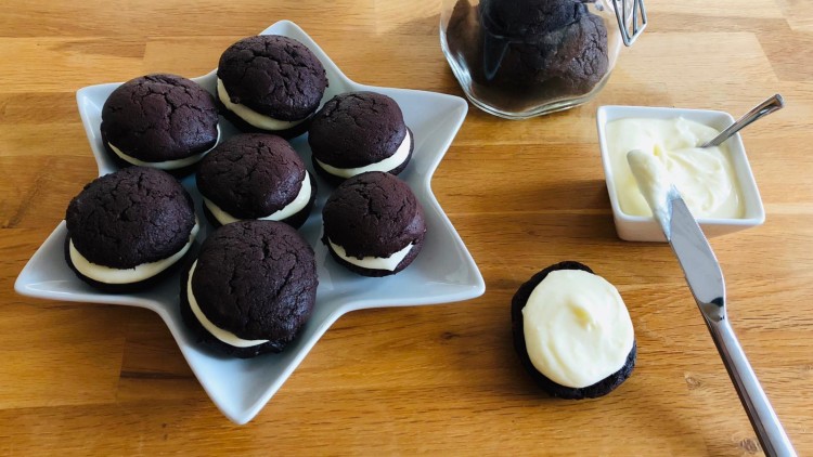 Acht dunkle Doppelkekse mit weißer Creme auf sternförmiger Platte, daneben ein Glas, Schälchen mit Creme und ein Messer.