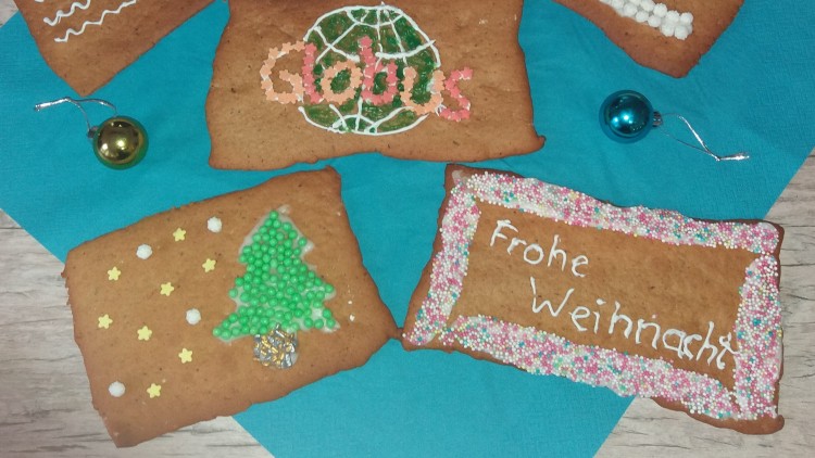 Fünf verzierte Lebkuchen mit Schriftzügen und Motiven wie Globus-Logo, Tanne und „Frohe Weihnacht“ liegen auf blauer Serviette.