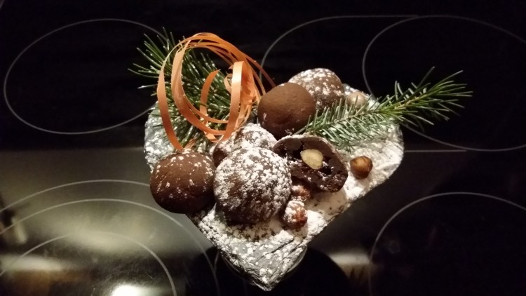 Herzförmige Schale mit Schokokugeln, Puderzucker, Haselnüssen, Tannenzweigen und orangem Band auf dunklem Untergrund.