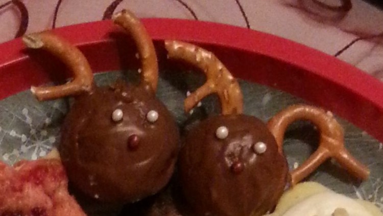 Zwei Rentier-Pralinen aus Schokolade mit Salzbrezel-Geweih, Zuckerperlen-Augen und rotem Zuckerdeko-Nase auf einem roten Teller.