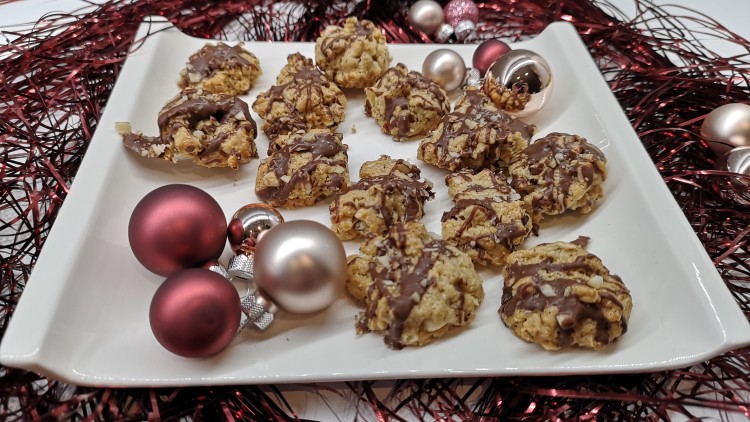 Plätzchen mit Schokoladenglasur liegen auf einem weißen Teller, dekoriert mit roten und silbernen Weihnachtskugeln sowie rotem Lametta.