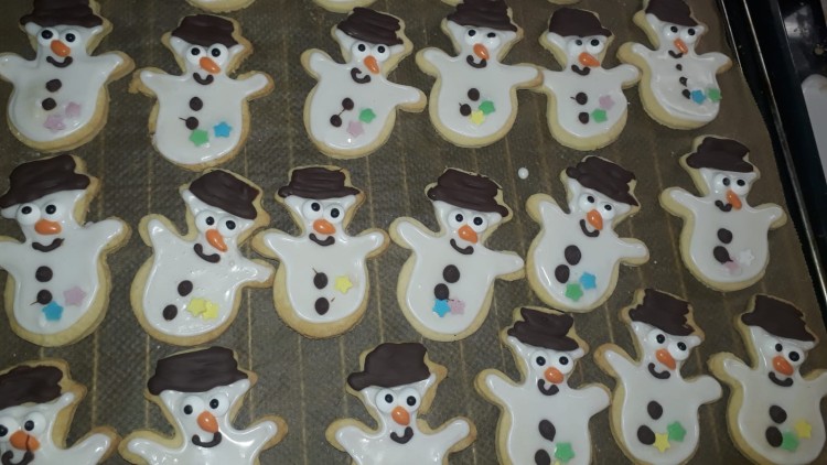 Mehrere Plätzchen in Schneemannform mit weißer Glasur, Schokohut, Möhrennase und bunten Sternen liegen auf einem Backblech.