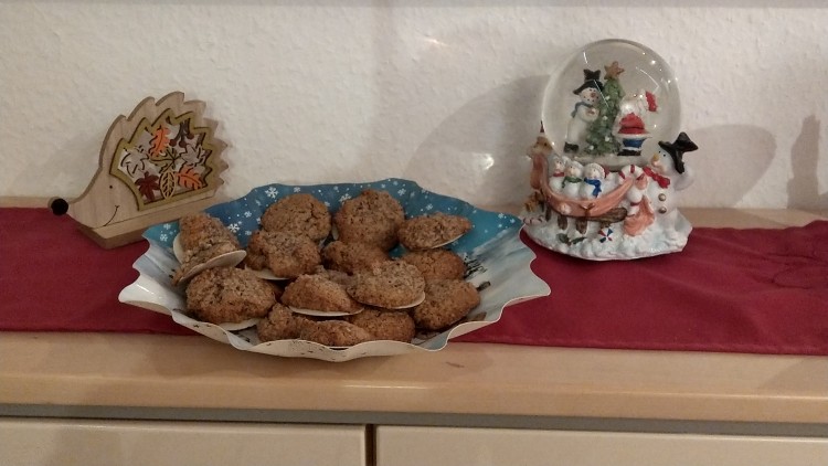 Ein Teller mit braunen Plätzchen, ein Holz-Igel mit Herbstdekoration und eine Schneekugel stehen auf einem roten Tuch.