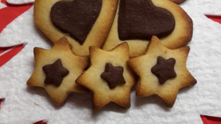 Herz- und sternförmige Kekse mit dunkler Dekoration liegen auf einem weißen Untergrund vor rotem Tuch mit Rentiermuster.